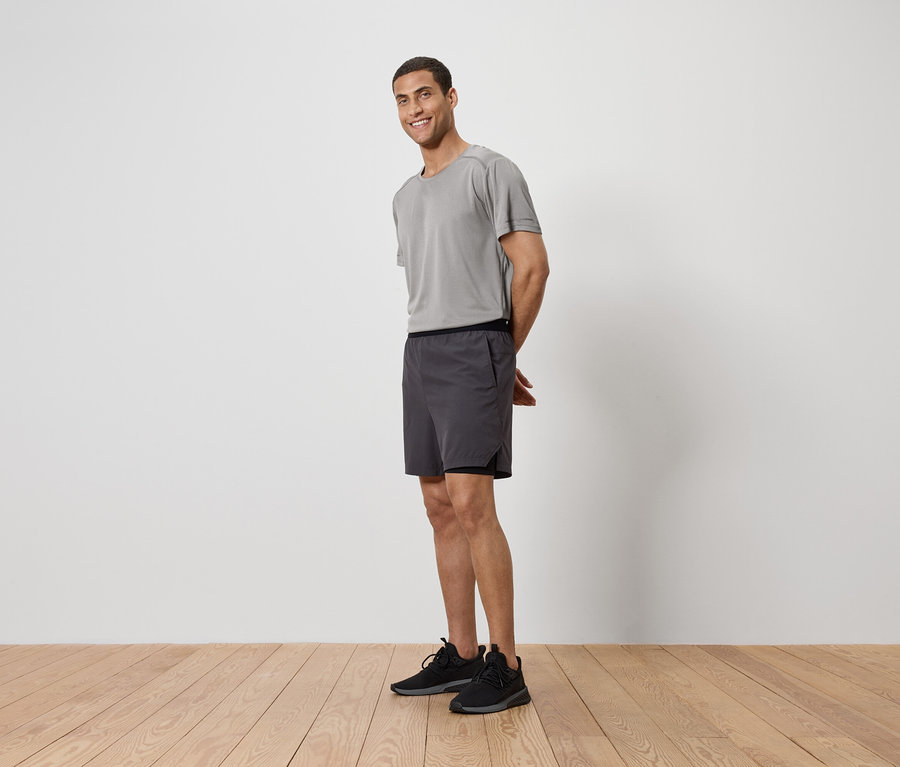 Mężczyzna w szarej koszulce, szortach i butach sportowych stoi na drewnianej podłodze przed białą ścianą.