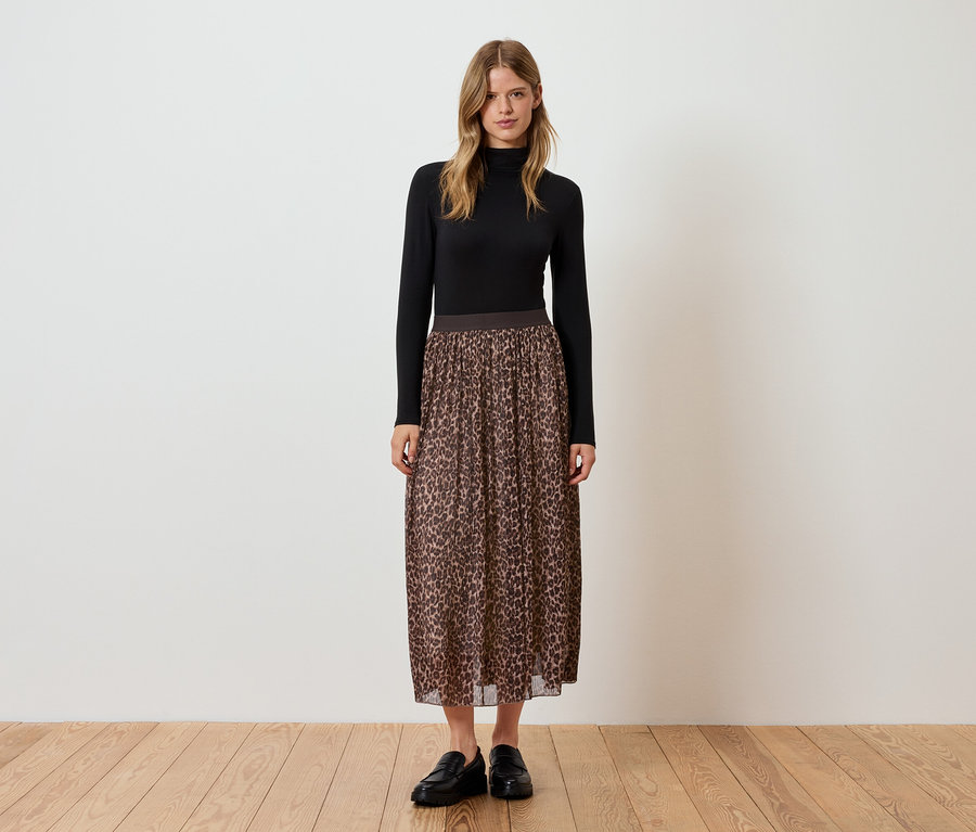 Kobieta stoi w czarnym golfie, brązowej plisowanej spódnicy we wzór zwierzęcy i czarnych butach.