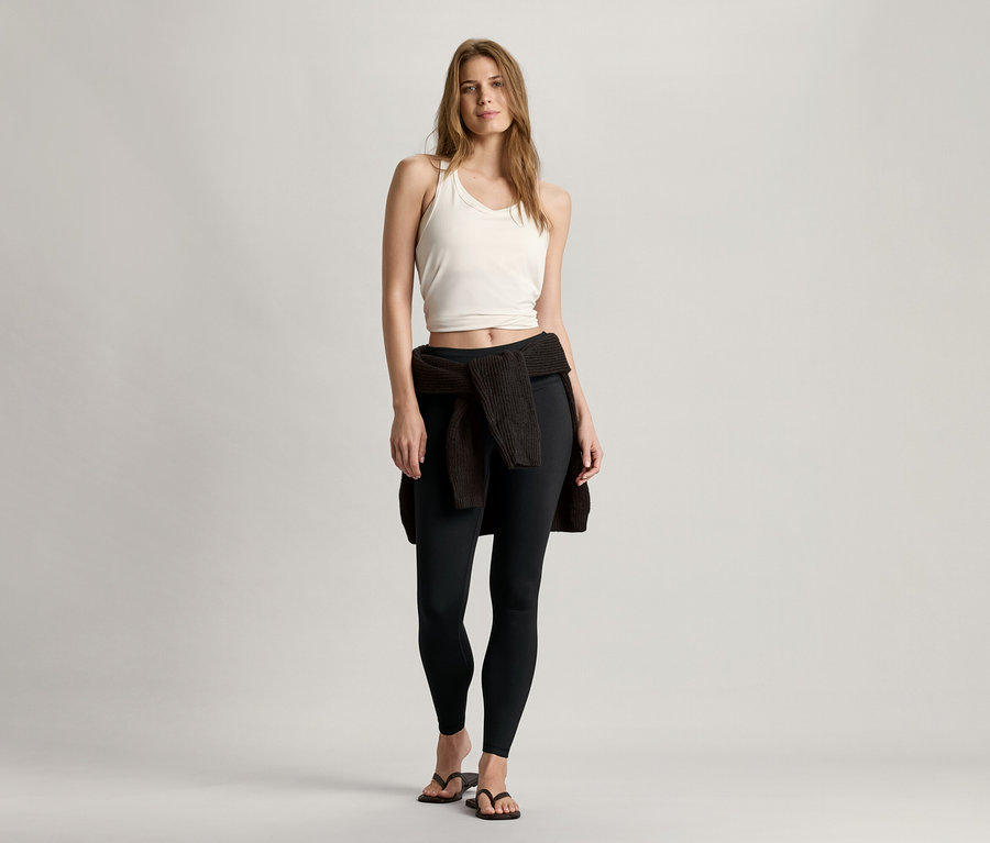 Kobieta stoi i ma na sobie białą bezszwową braletkę sportową, czarne legginsy i sweter zawiązany w pasie.