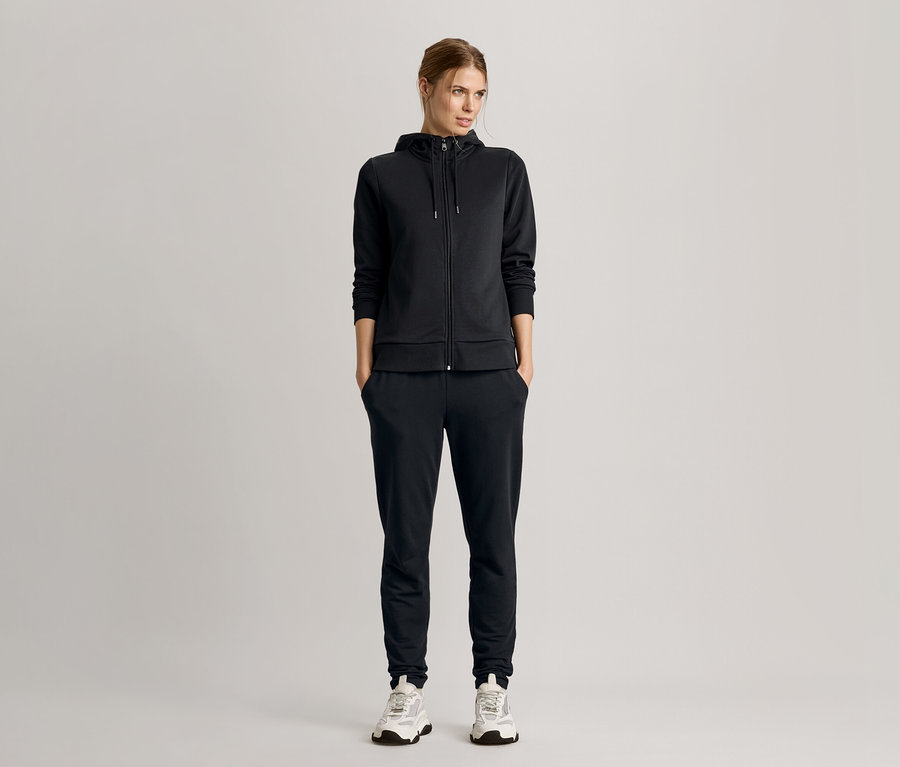 Kobieta stoi z rękami w kieszeniach, ubrana w czarny dres z bluzą z kapturem i trampki.