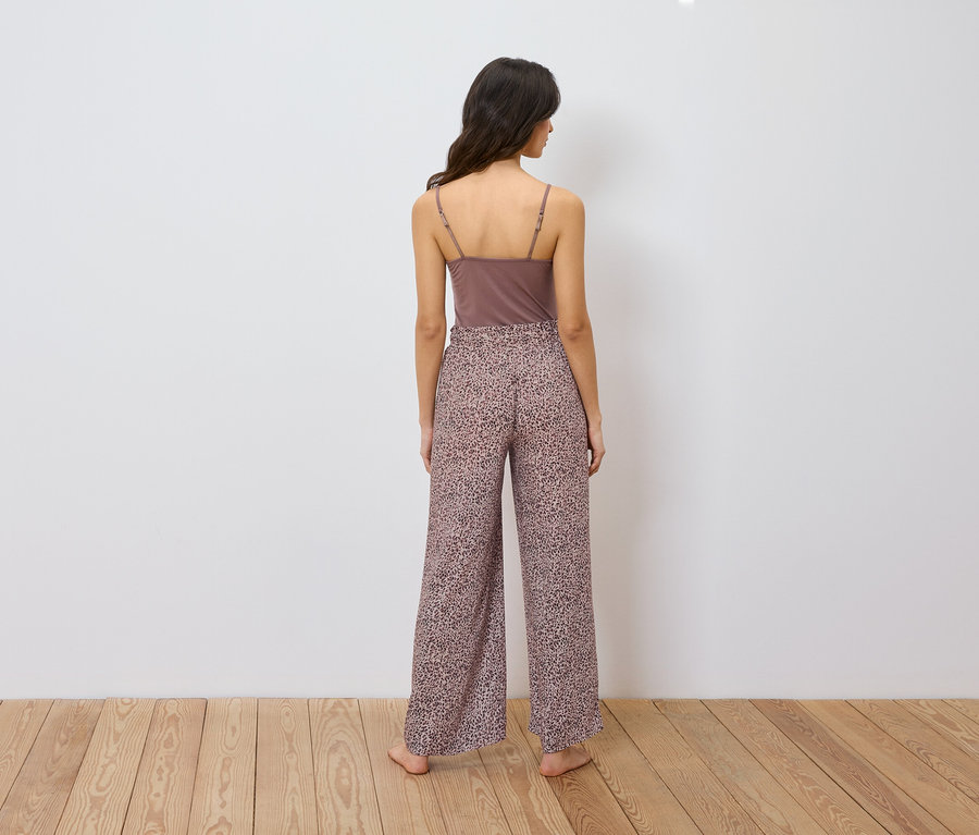 Kobieta stoi tyłem, ubrana w brązowy podkoszulek na ramiączkach i szerokie spodnie we wzór zwierzęcy.