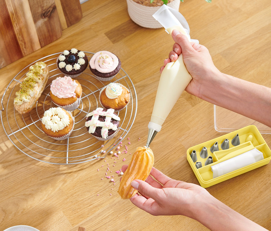 Osoba dekoruje ciastko za pomocą worka cukierniczego, babeczki i żółte pudełko z tylkami leżą na drewnianym stole.