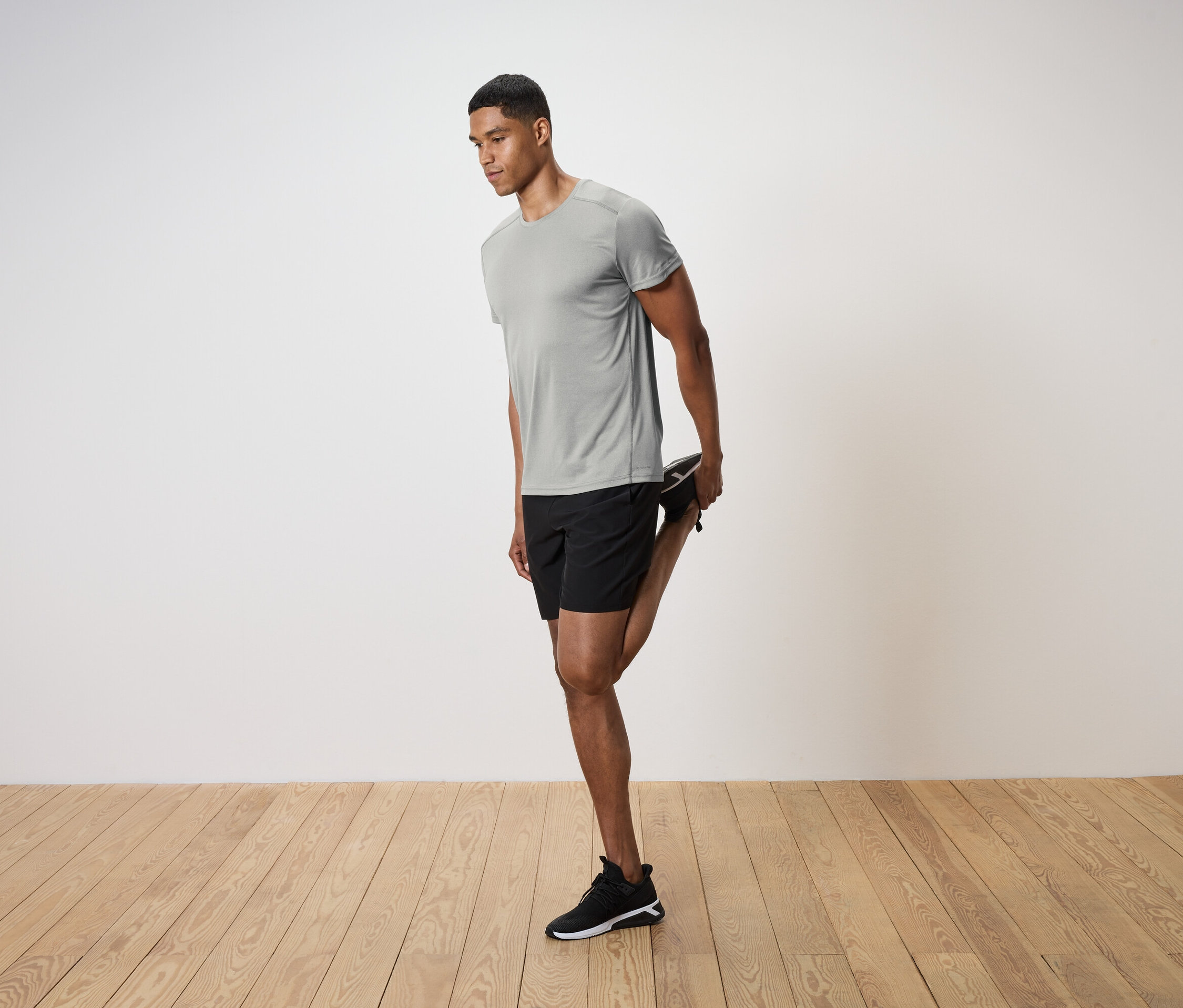 Mężczyzna w szarej koszulce i czarnych szortach rozciąga nogę na drewnianej podłodze.