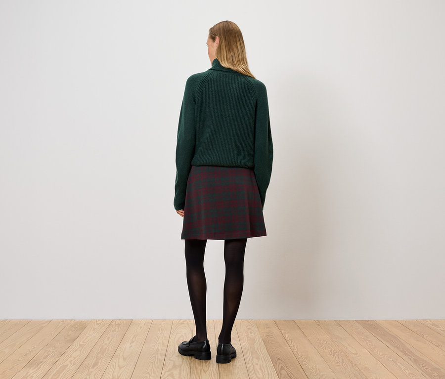 Kobieta stoi tyłem, ubrana w zielony sweter, spódnicę w kratę i czarne rajstopy.