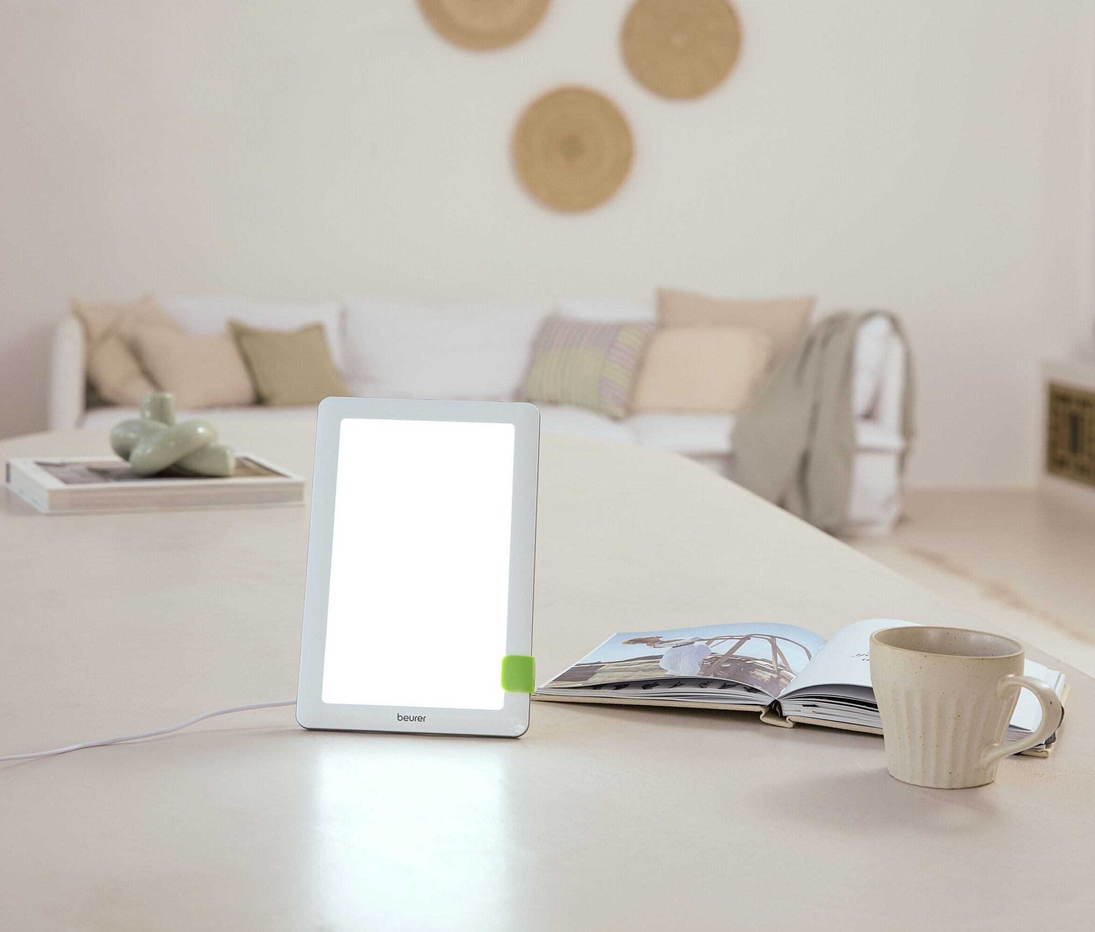 Na stole stoi lampa, otwarta książka i kubek. W tle znajduje się sofa.