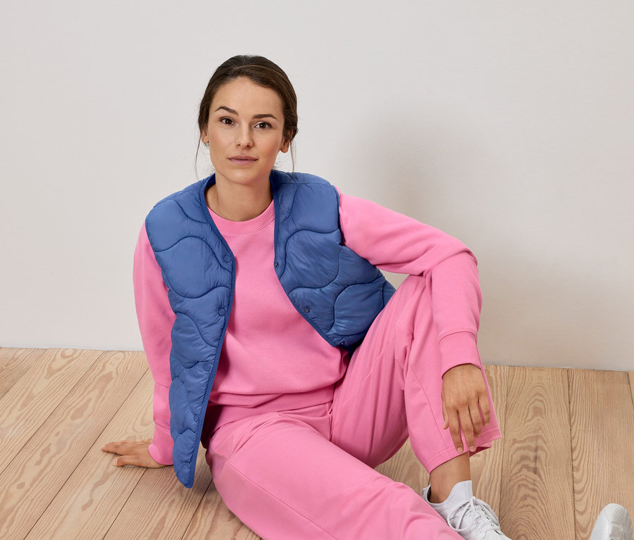 Kobieta siedzi na podłodze w różowym dresie i niebieskiej kamizelce.