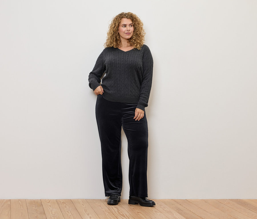 Kobieta o kręconych włosach pozuje w szarym swetrze w warkocze i czarnych welurowych spodniach.