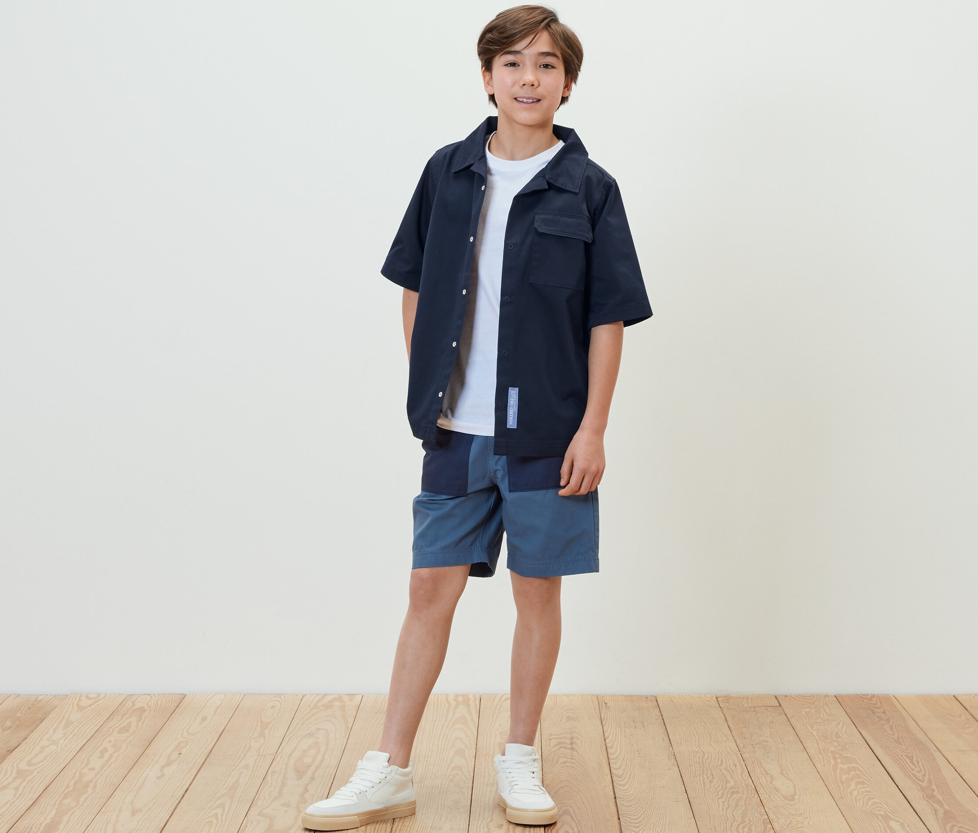 Chłopiec stoi w koszuli, szortach i tenisówkach.