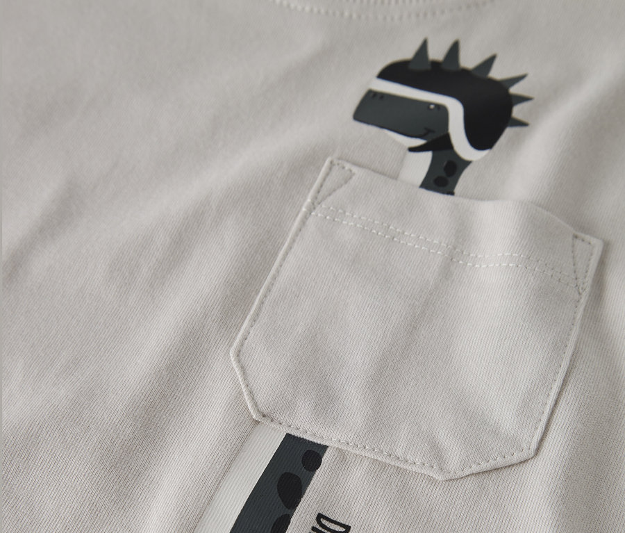 Zbliżenie na jasno szary T-shirt z nadrukowaną postacią dinozaura w kasku, wystającą z kieszeni na piersi. Na T-shircie widnieje napis "Dino Racer Born to Race".
