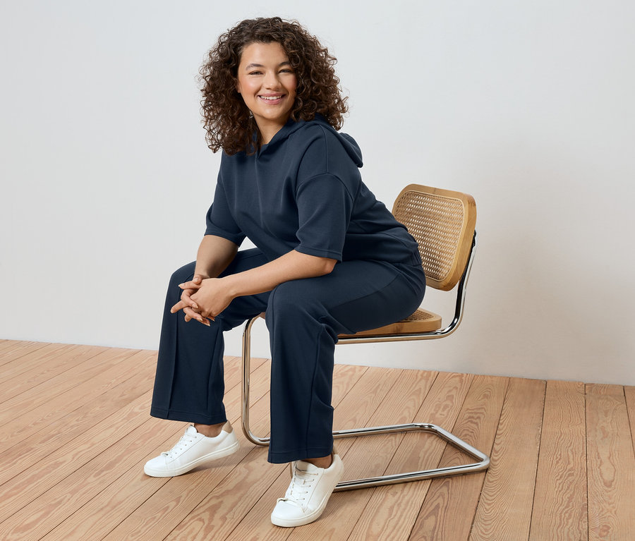 Kobieta o kręconych włosach siedzi na krześle, ubrana w granatowy dres i białe tenisówki.