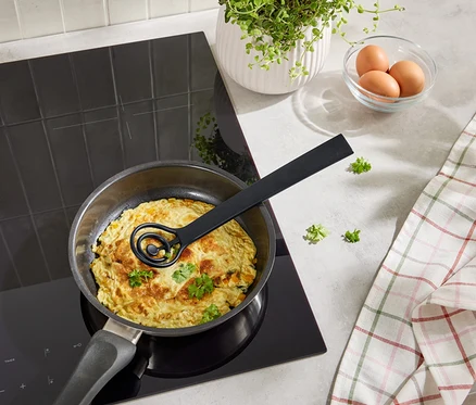 Omlet gotowany jest na czarnej płycie grzejnej na patelni z czarną trzepaczką. Trzy jajka w misce, roślina, ręcznik kuchenny.