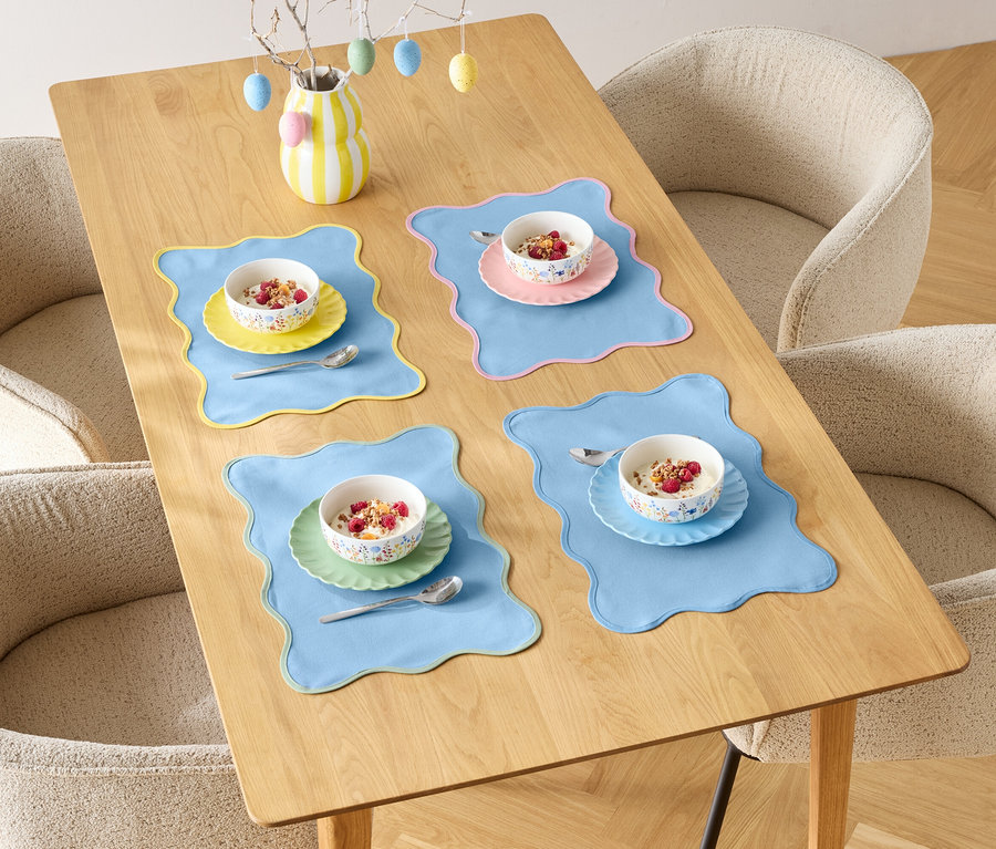 Stół jadalny nakryty dla czterech osób z miskami płatków śniadaniowych i kolorowymi podkładkami, otoczony czterema krzesłami.