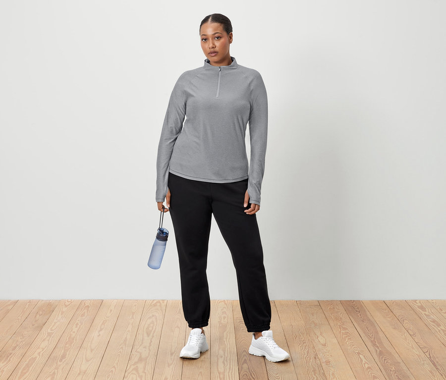 Kobieta stoi na tle białej ściany, trzymając w ręku niebieską butelkę z wodą. Ma na sobie szarą koszulę z długim rękawem, czarne spodnie dresowe i białe tenisówki.