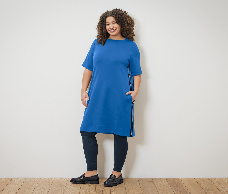 Kobieta o kręconych włosach w niebieskiej sukience z zamkiem błyskawicznym i legginsach pozuje na tle ściany.