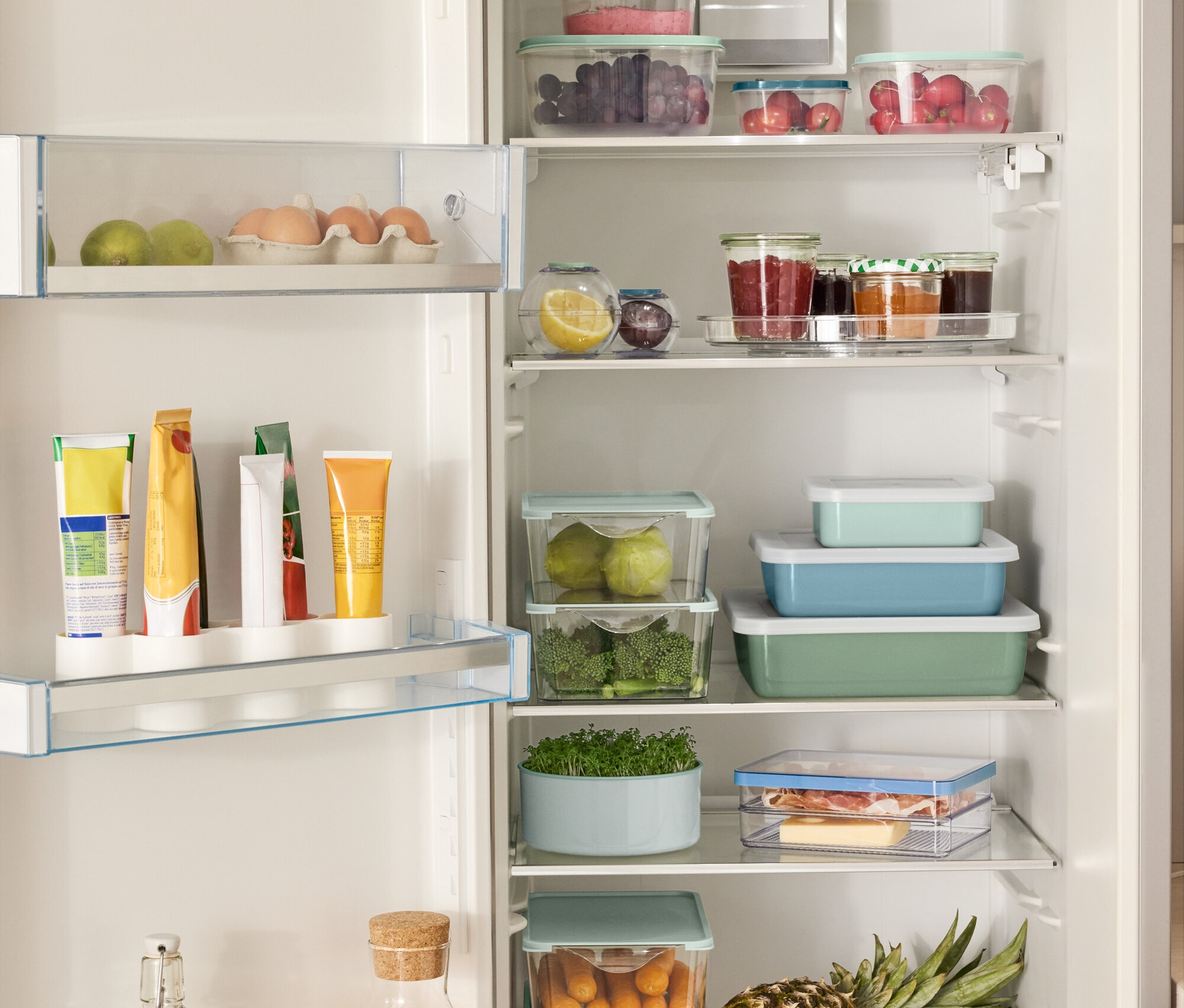 Otwarta lodówka wypełniona różnymi produktami spożywczymi, w tym 4 pudełka do przechowywania, 2 pojemniki spożywcze w kształcie kuli, 3 emaliowane formy do zapiekania z pokrywką i obracany talerz.