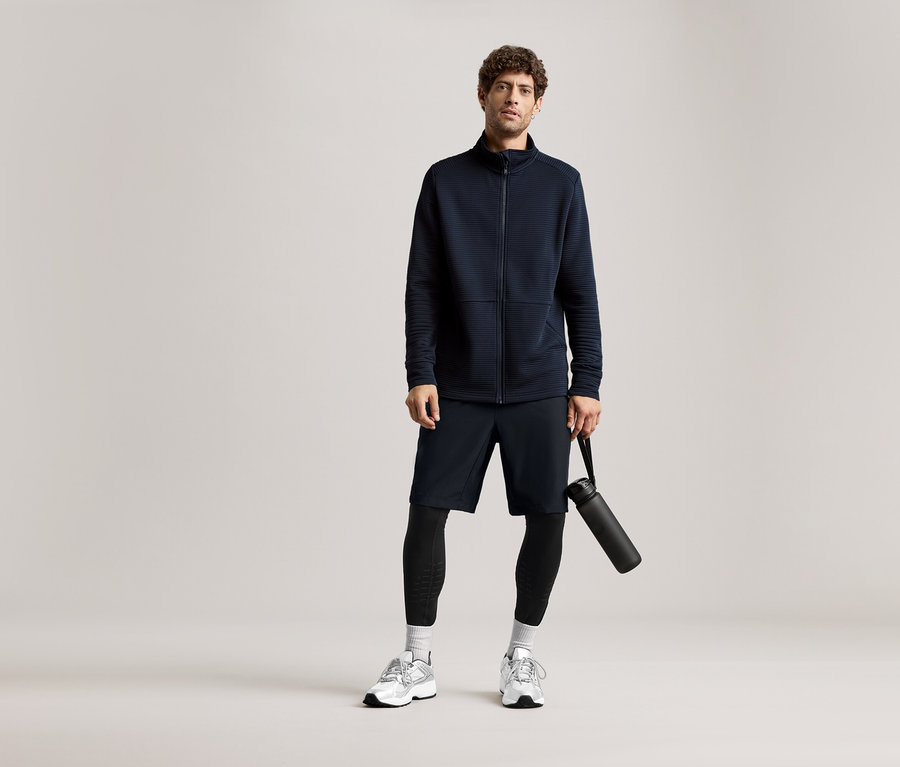 Mężczyzna w granatowej kurtce, szortach i legginsach trzyma czarną butelkę na wodę.