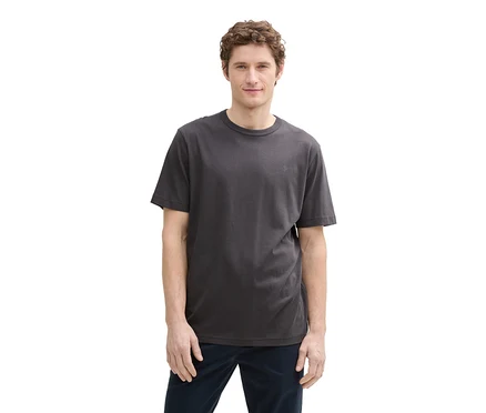 Mężczyzna z kręconymi włosami w ciemnoszarym T-shircie Tom Tailor z logo i ciemnoniebieskich spodniach.