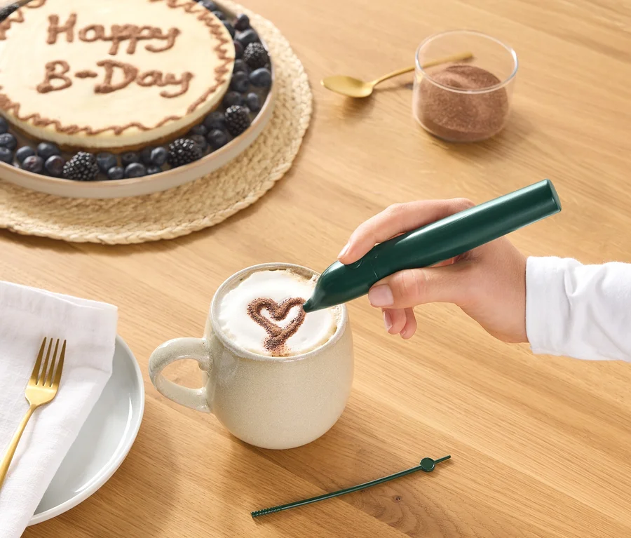 Ręka rysuje zielonym pisakiem do dekoracji kakaowe serce na piance kawy w kubku. Na drewnianym stole widoczne są również tort z napisem „Happy B-Day” i jagodami, szklanka kakao w proszku, złota łyżeczka oraz talerz z serwetką i widelcem.