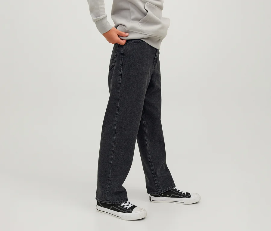 Osoba w czarnych jeansach Jack&Jones JJOrginial Alex, jasnoszarej bluzie z kapturem i czarno-białych sneakersach, z jedną ręką trzymającą pas jeansów.