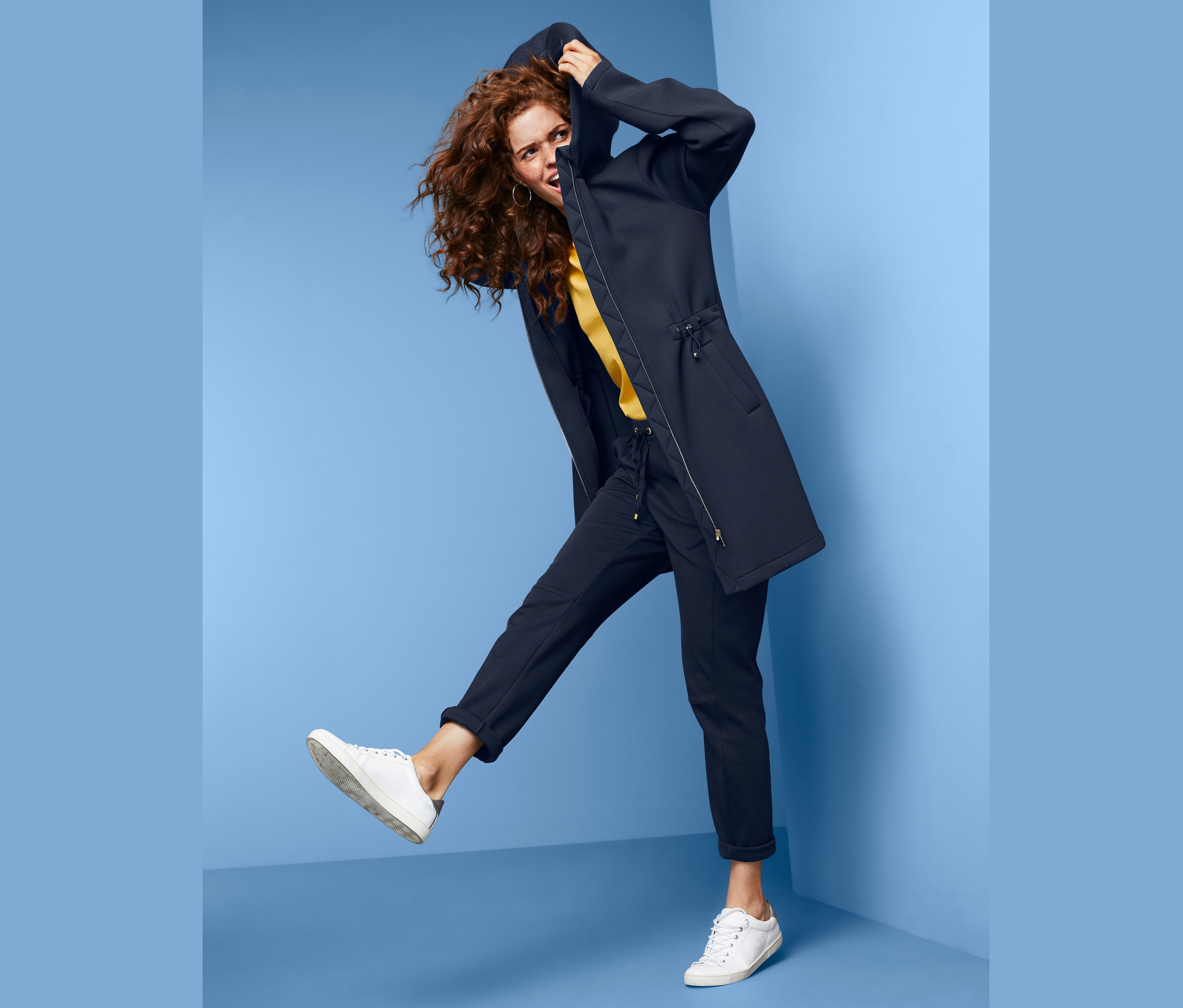 Kobieta o kręconych włosach pozuje w płaszczu dresowym z kapturem na niebieskim tle.