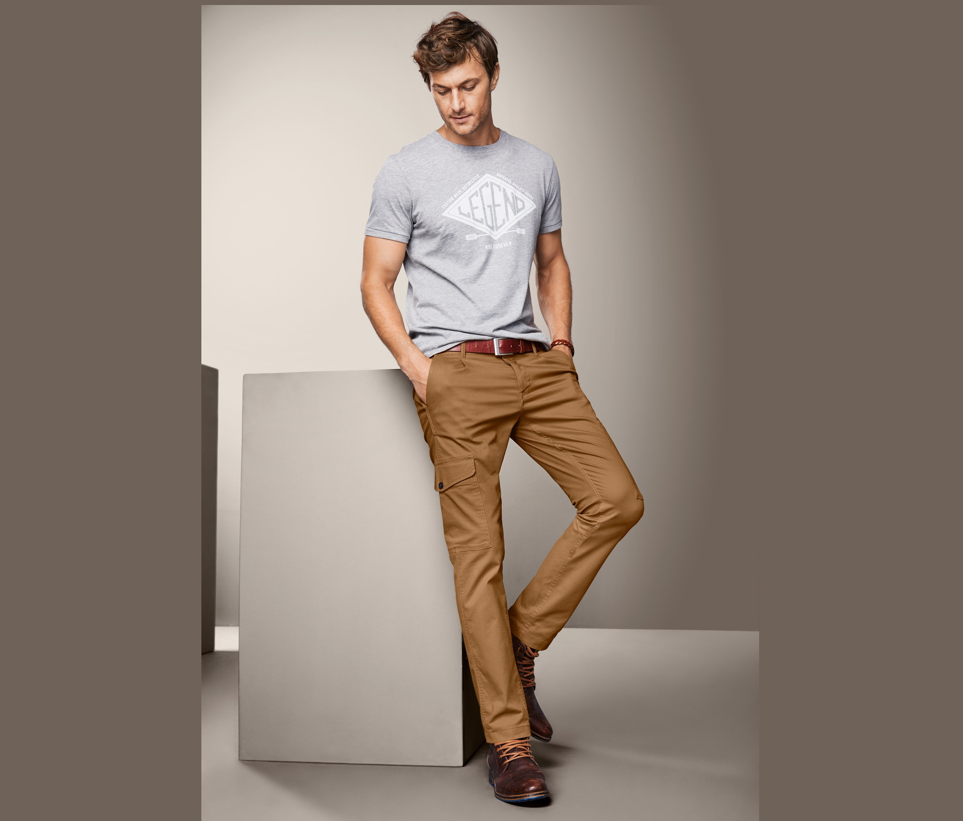 Mężczyzna w szarym t-shircie i męskich spodniach bojówkach opiera się o szary sześcian.