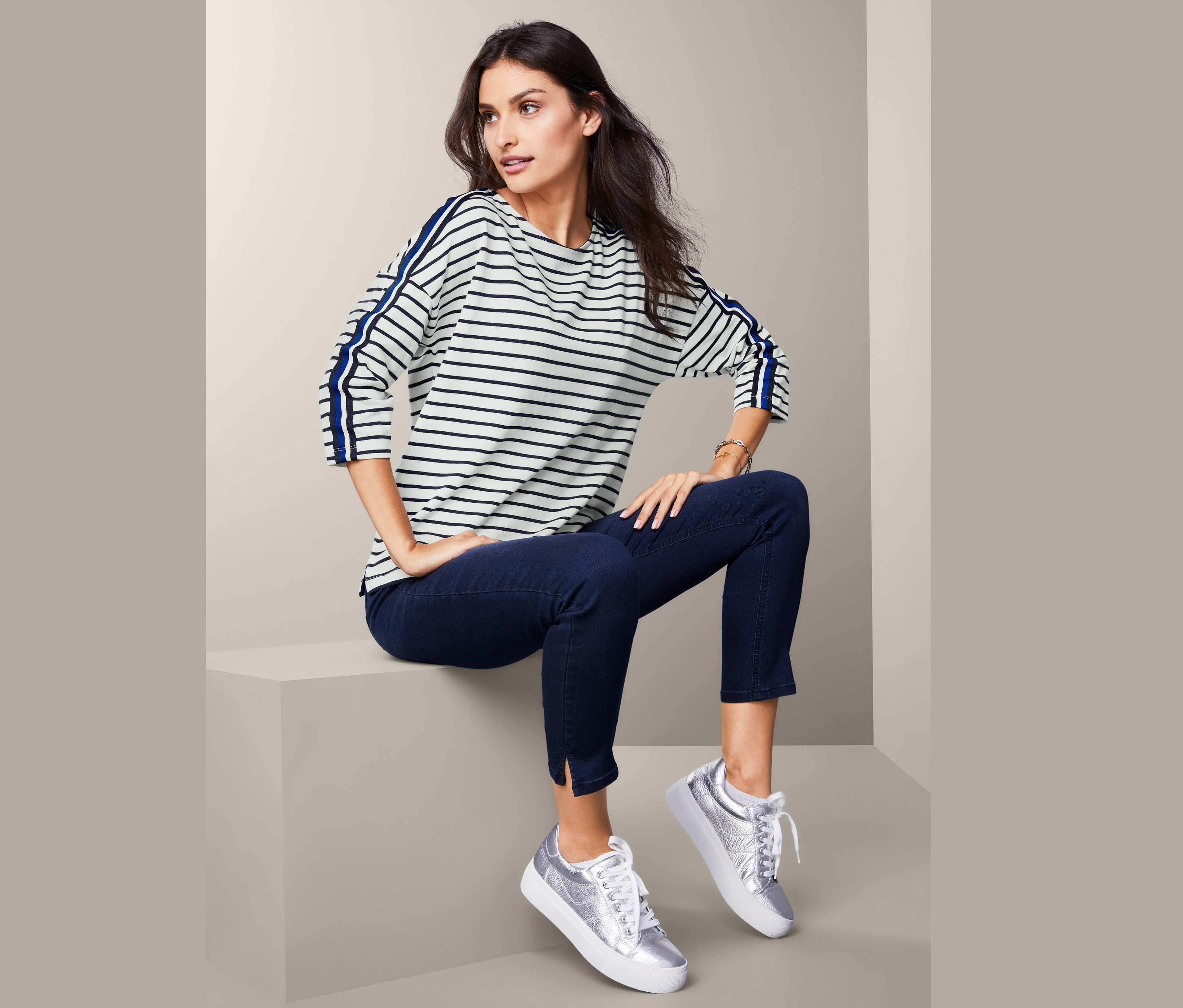 Kobieta siedzi i ma na sobie bluzę z nietoperzowymi rękawami z niebieskimi paskami na rękawach, ciemne dżinsy i srebrne tenisówki.