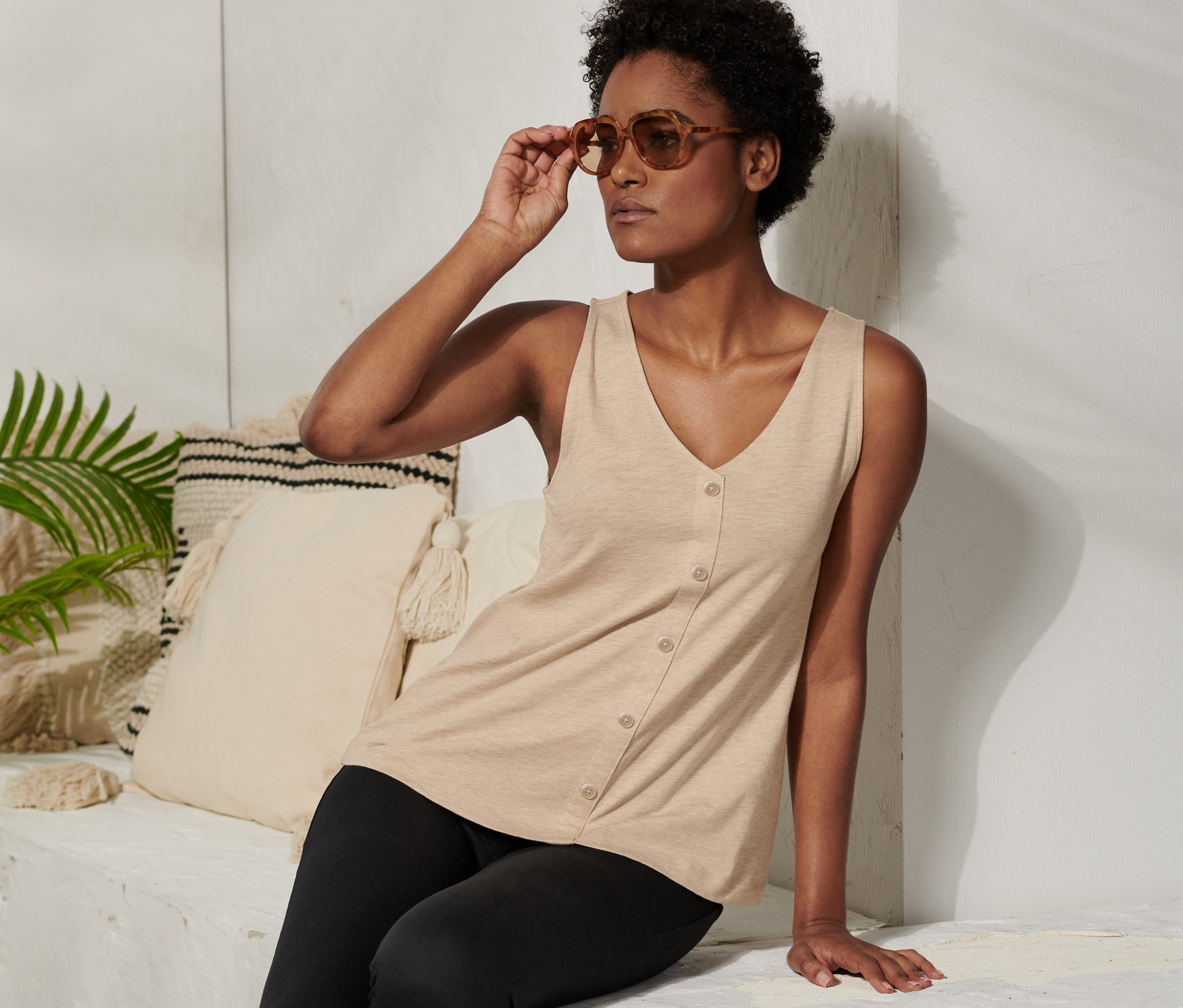 Kobieta siedzi i poprawia okulary przeciwsłoneczne. Ma na sobie beżową koszulkę z plisą z guzikami i czarne legginsy.