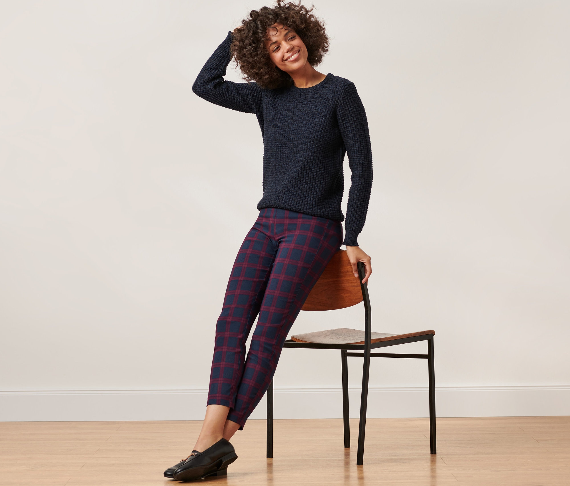 Kobieta siedzi na krześle, ma na sobie niebieski sweter i czerwono-niebieskie spodnie stretchowe w kratkę.