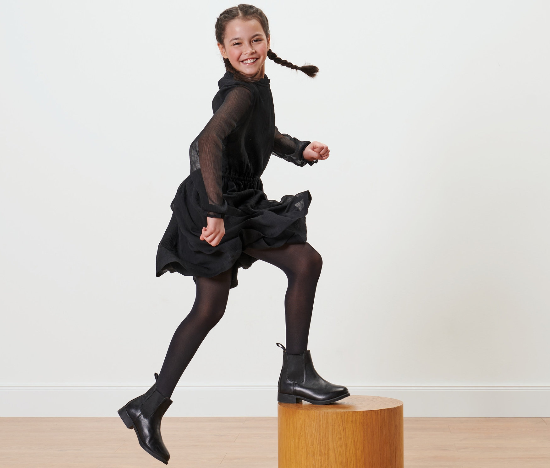Uśmiechnięta dziewczyna w czarnej sukience i czarnych rajstopach nylonowych, 2 pary, skacze z drewnianego cokołu.
