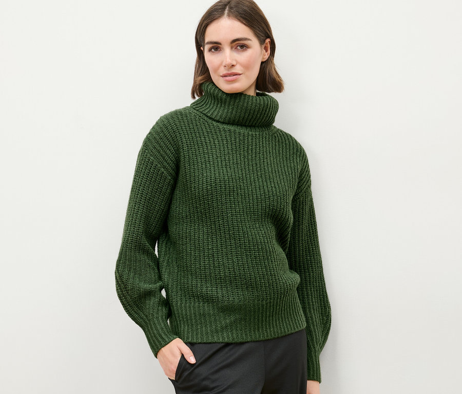 Kobieta pozuje na jasnym tle w swetrze o grubym splocie z golfem.