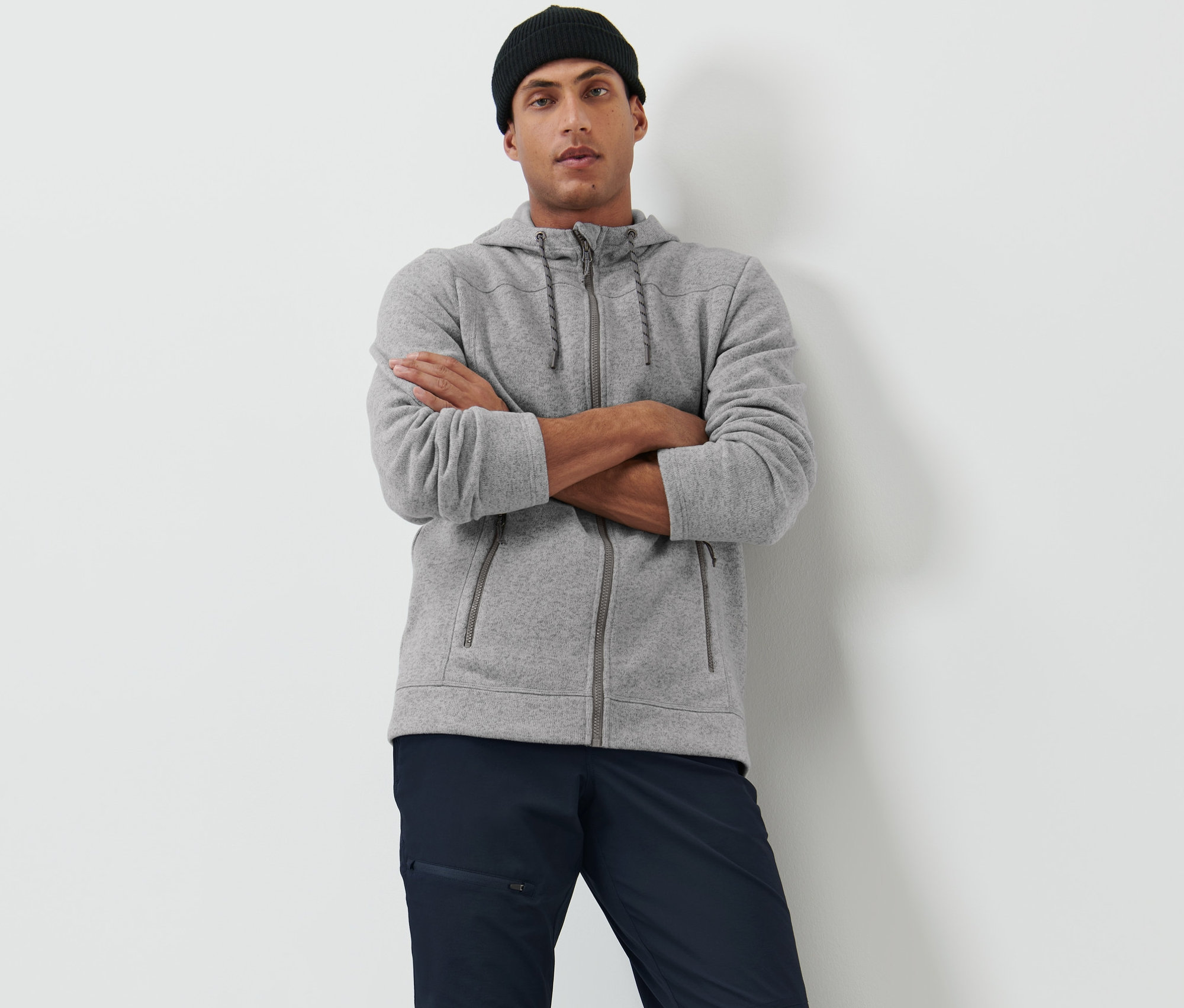 Mężczyzna pozuje ze skrzyżowanymi ramionami w szarej rozpinanej bluzie z dzianiny polarowej i czarnej czapce.