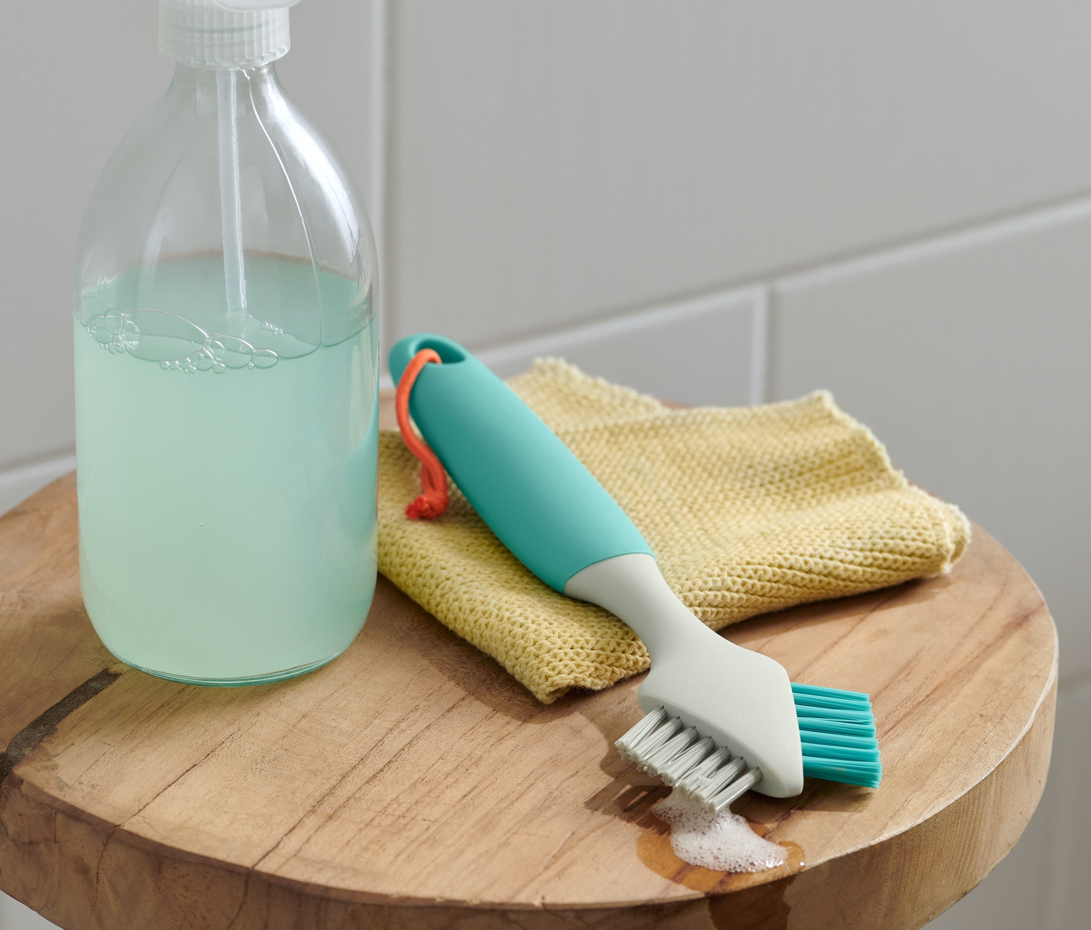 Szczotka do fug, miękka/twarda leży na żółtej ściereczce obok środka czyszczącego.