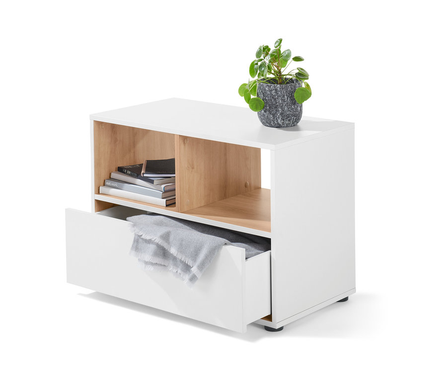 Biała szafka z szufladą, na której stoi roślina w szarej doniczce. W otwartej przegródce leżą książki, a w szufladzie szary koc.