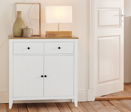 W pokoju stoi komoda z szufladami, szafka typu sideboard, regał i lampa stołowa z drewnianą podstawą.