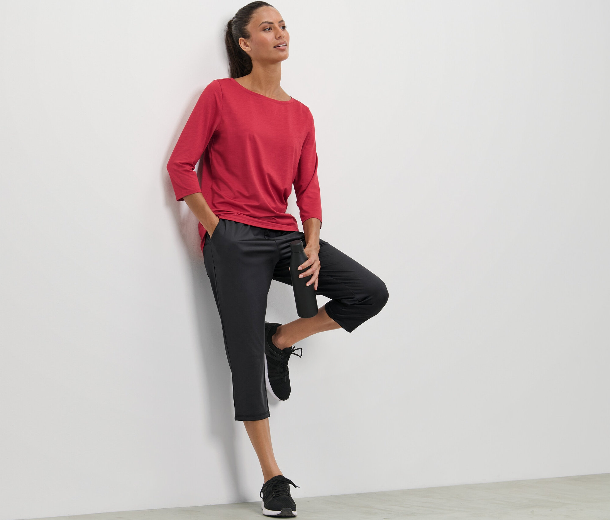 Kobieta opiera się o ścianę, ubrana w czerwoną koszulkę i spodnie sportowe o długości 3/4.