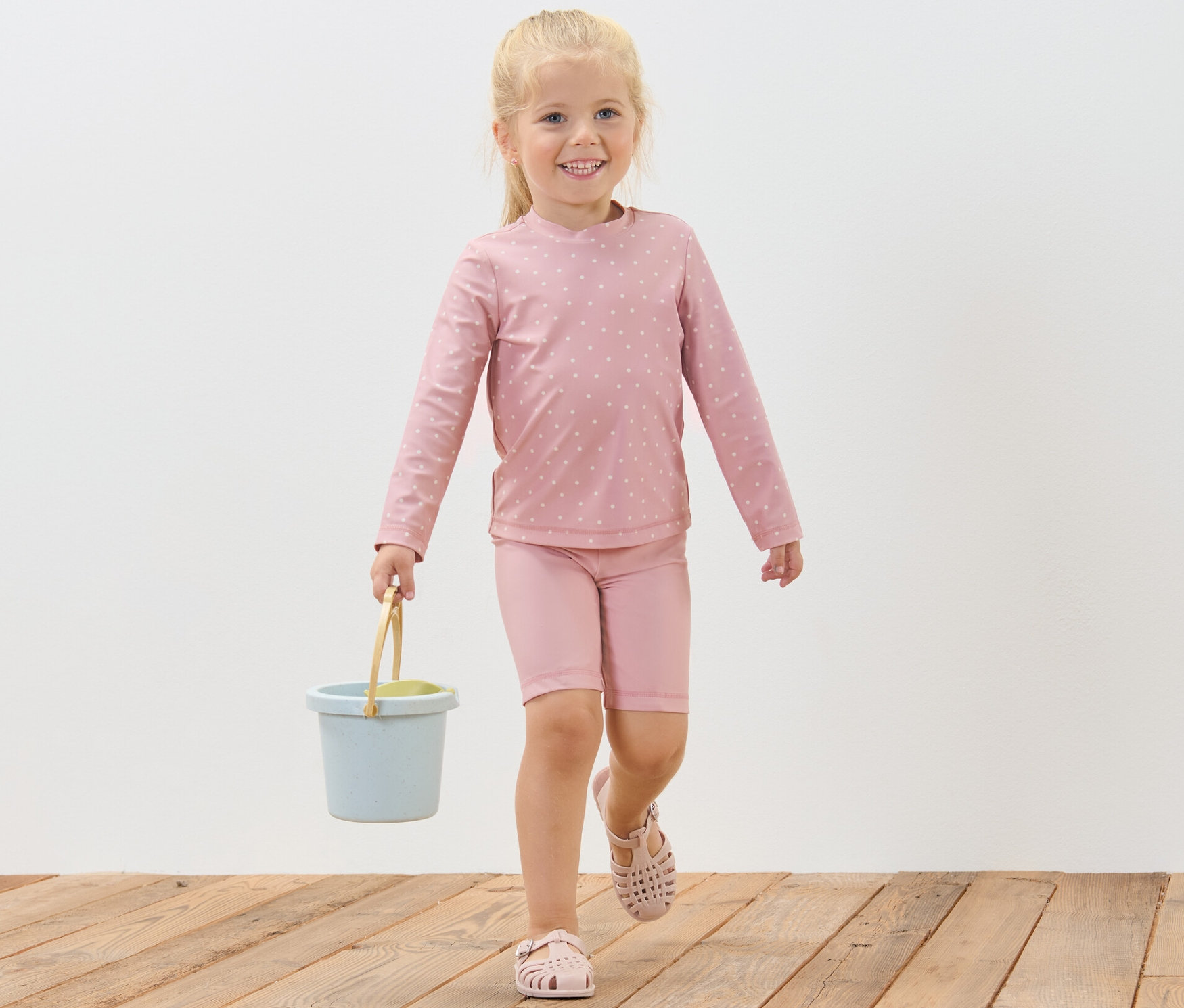 Uśmiechnięta dziewczynka idzie po drewnianej podłodze, niosąc niebieskie wiadro. Ma na sobie komplet dziecięcy z filtrem UV 80, w kropki.
