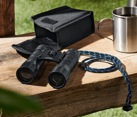 Na drewnianej ławce leżą lornetka kieszonkowa »12x32«, etui, metalowy kubek i butelka.