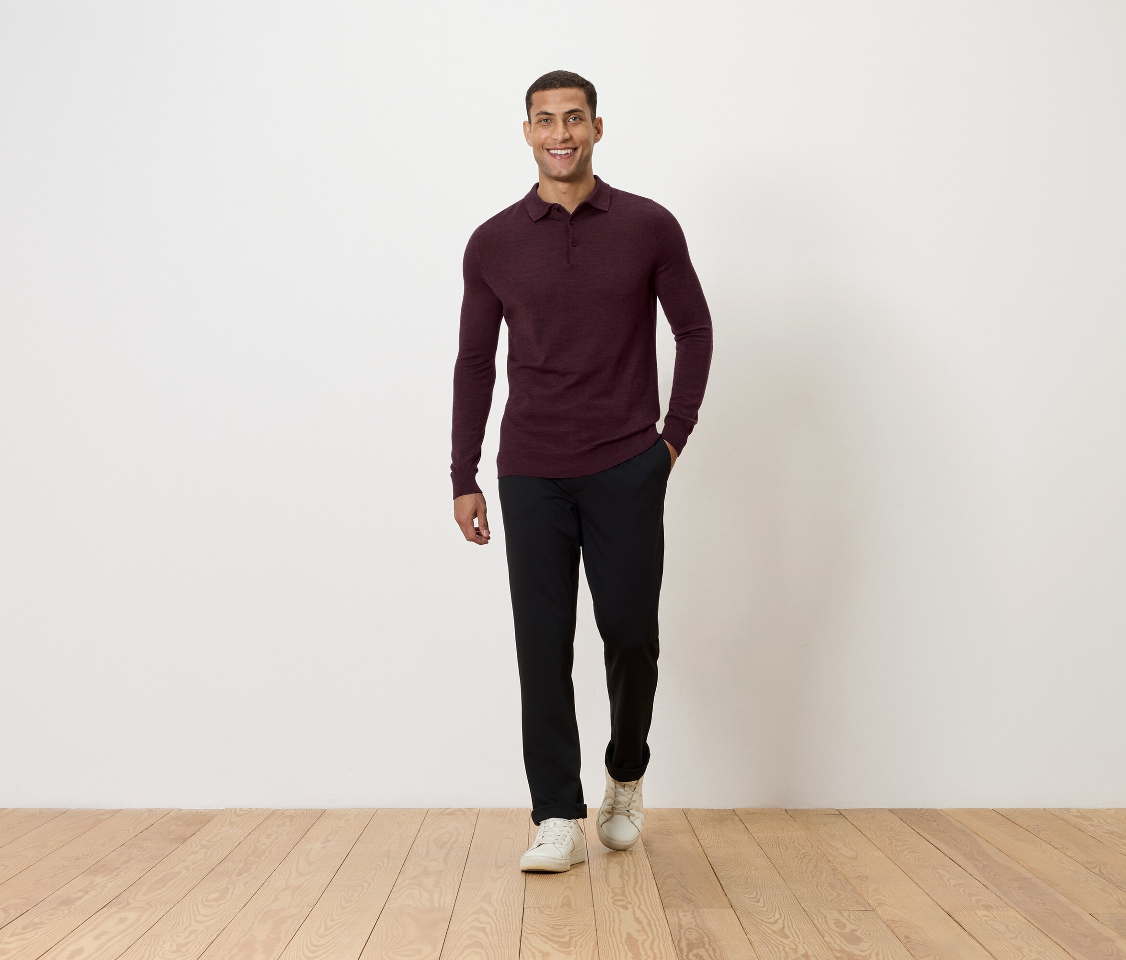 Mężczyzna idzie po drewnianej podłodze. Ma na sobie bordową koszulkę polo z długim rękawem, czarne spodnie i białe tenisówki.