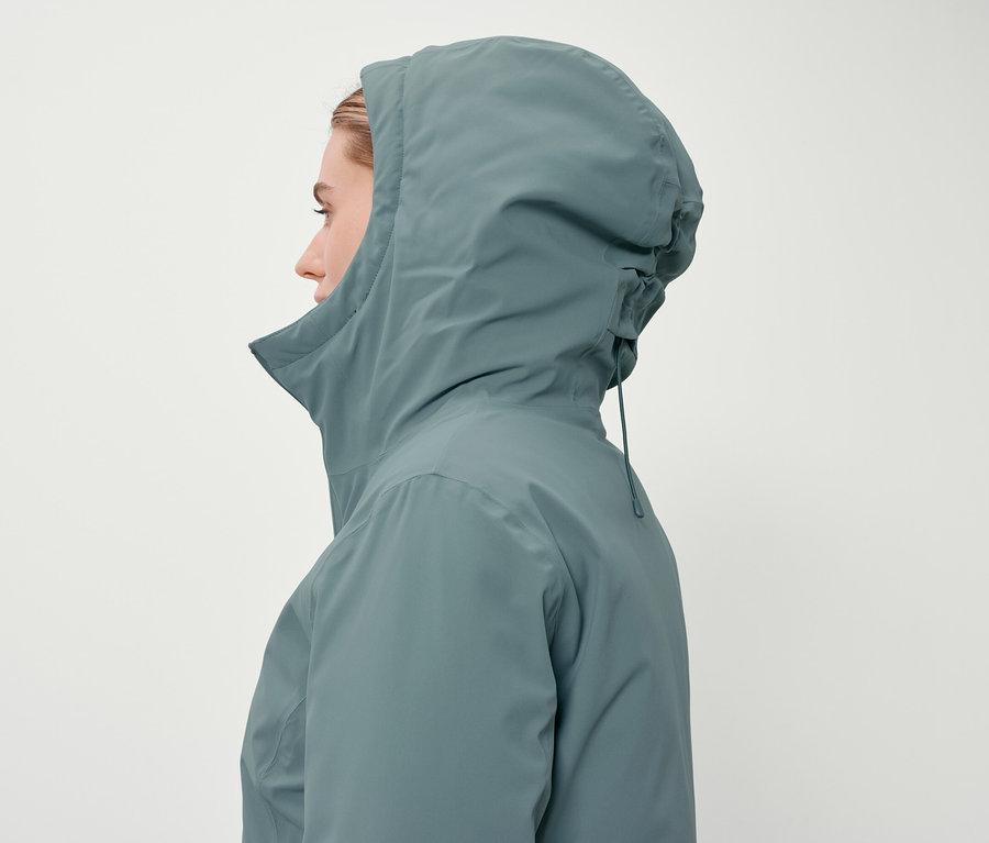 Profil kobiety w kapturze w niebiesko-zielonej kurtce na białym tle.