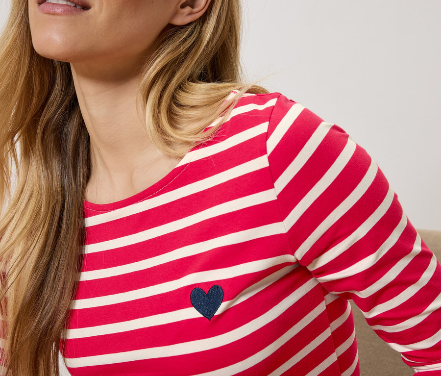 Blondwłosa kobieta ma na sobie czerwono-białą koszulkę w paski z długim rękawem i granatowym sercem.