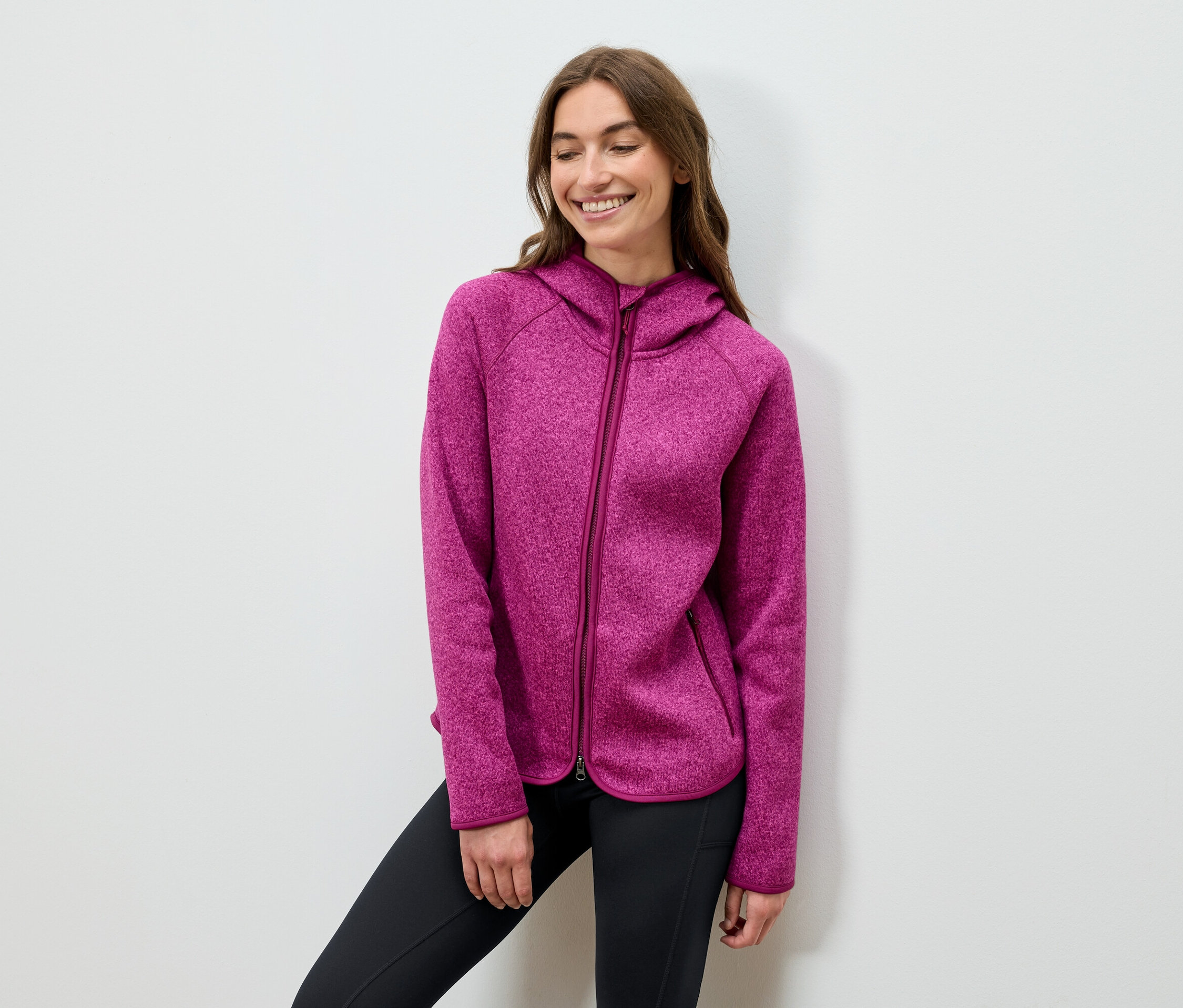 Uśmiechnięta kobieta pozuje w różowej bluzie z kapturem i czarnych spodniach.