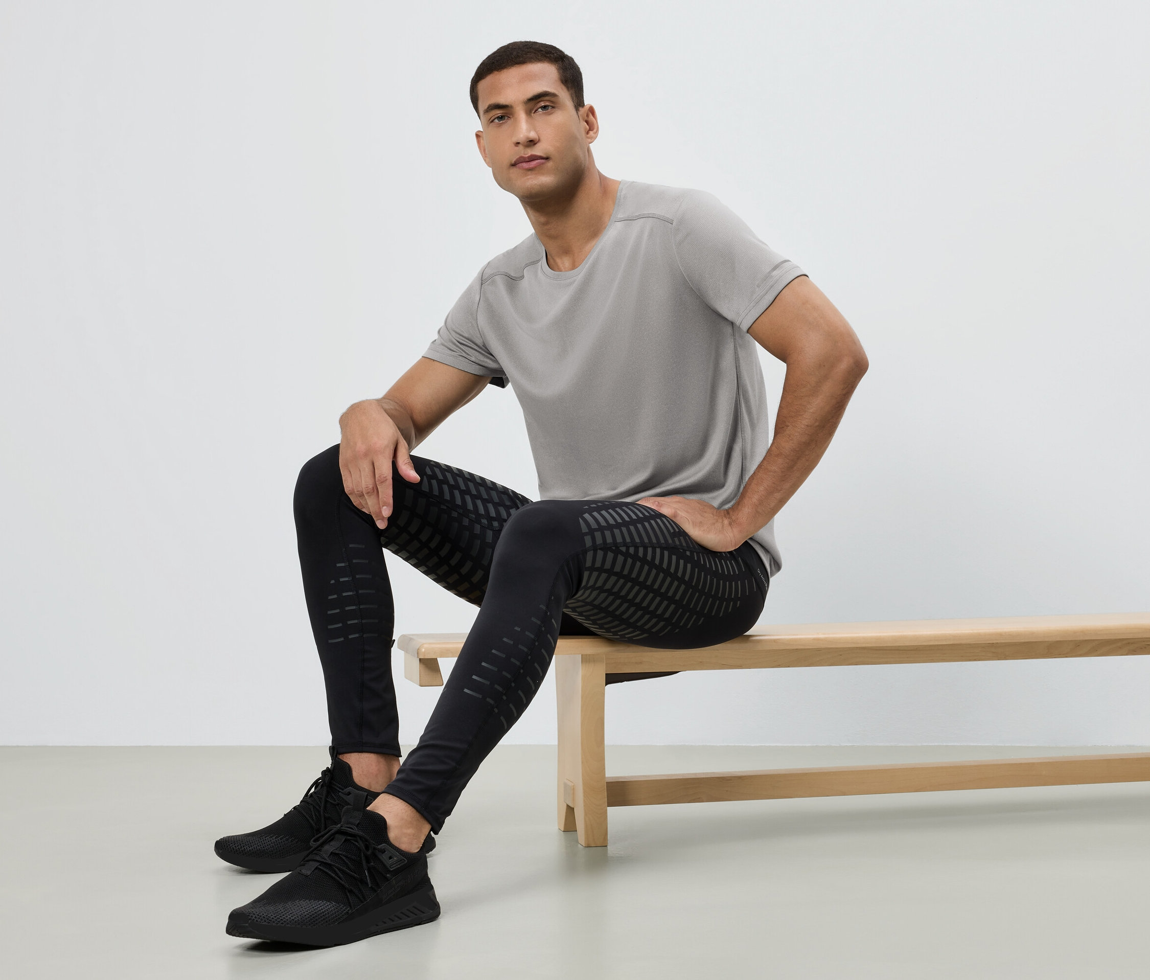 Mężczyzna siedzi na ławce, ubrany w szarą koszulkę, czarne legginsy we wzory i czarne buty sportowe.