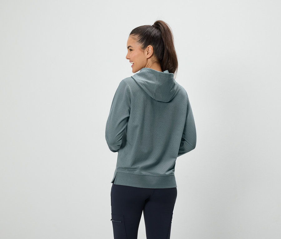 Kobieta z brązowymi włosami związanymi w koński ogon stoi tyłem, ubrana w niebiesko-zieloną bluzę z kapturem i granatowe spodnie.