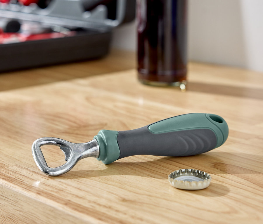 Na drewnianej powierzchni leży otwieracz do butelek obok kapsla, w tle butelka i zestaw narzędzi.