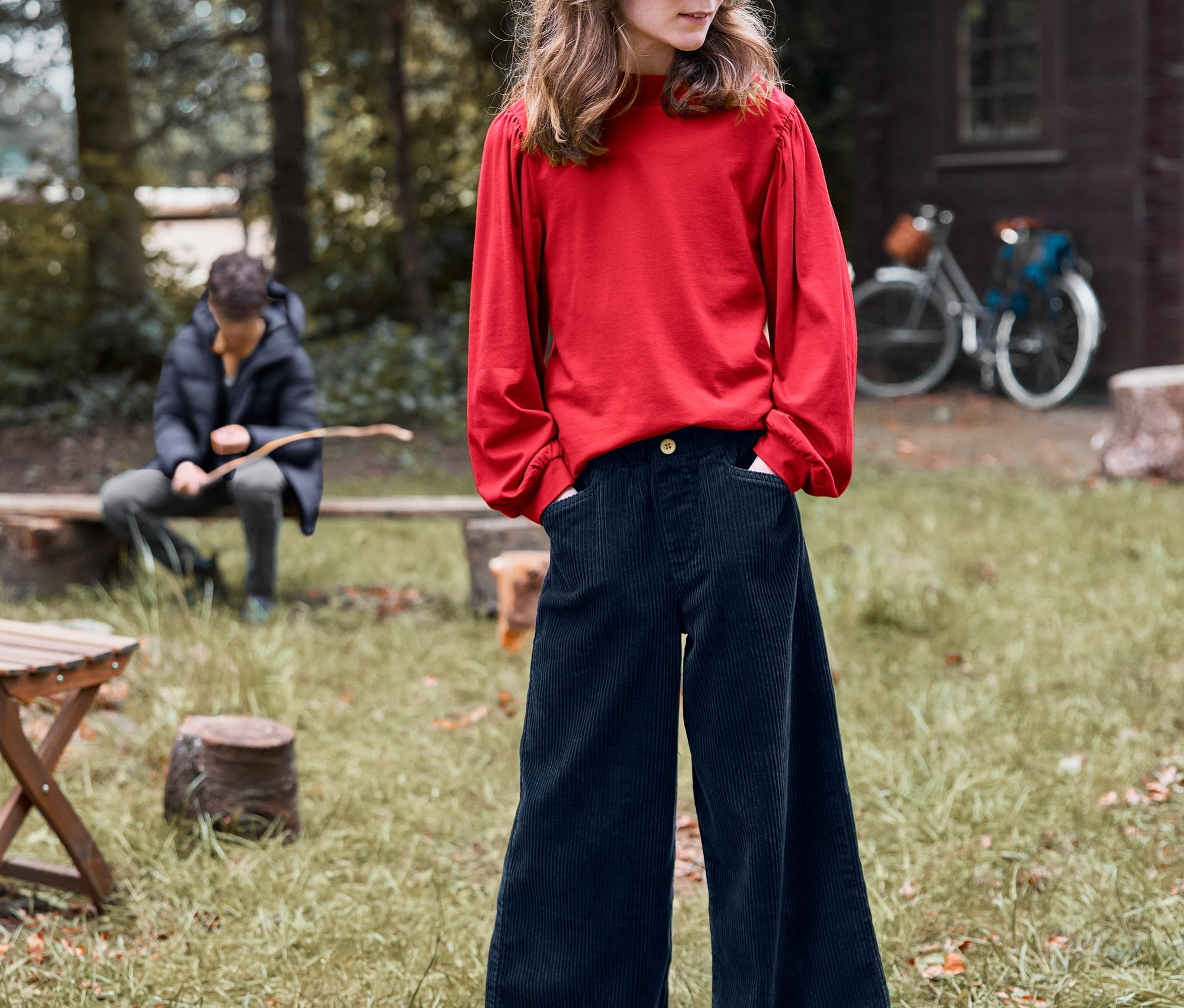 Dziewczynka stoi na zewnątrz z rękami w kieszeniach sztruksowych kulotów i bluzce z długim rękawem.