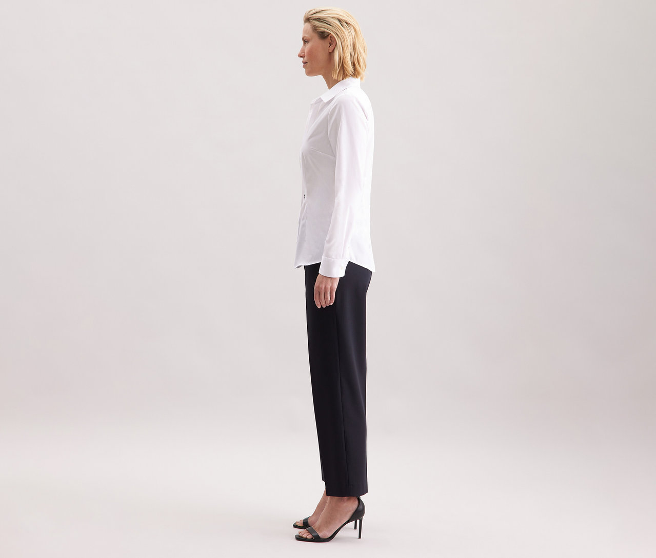 Profil kobiety noszącej białą bluzkę Seidensticker Slim Fit non iron, w połączeniu z ciemnymi spodniami i czarnymi butami na wysokim obcasie.