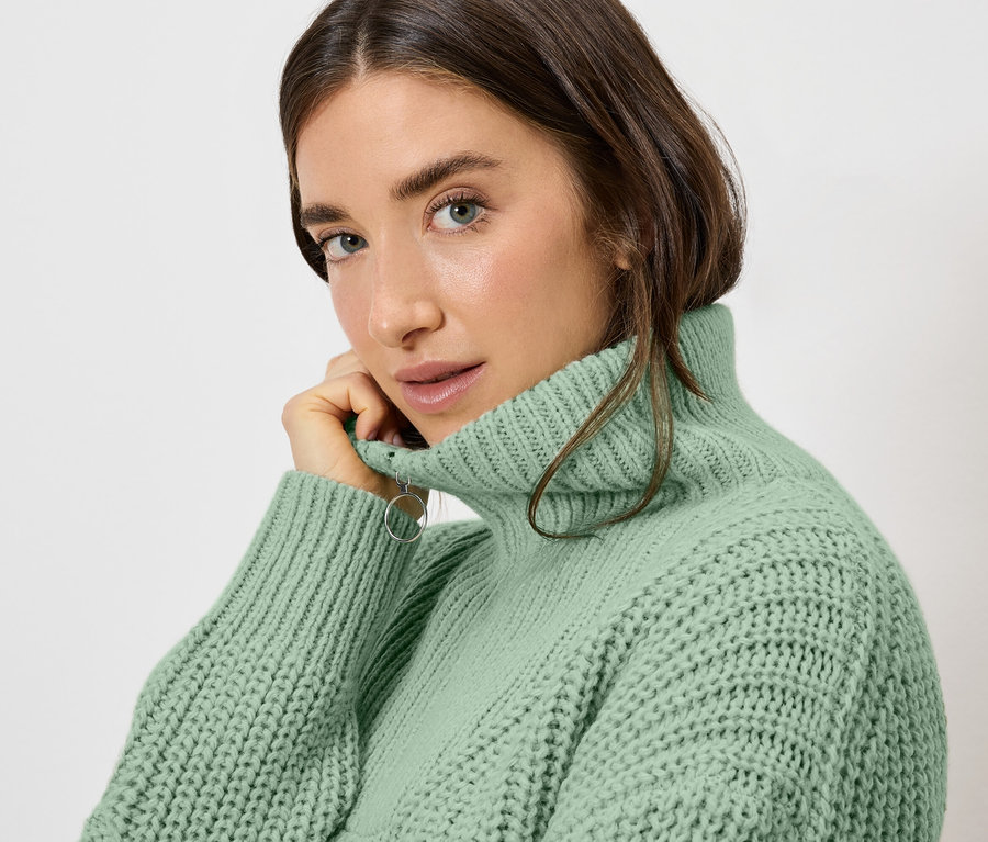 Kobieta o jasnobrązowych włosach w jasnozielonym golfie pozuje i patrzy w kamerę.