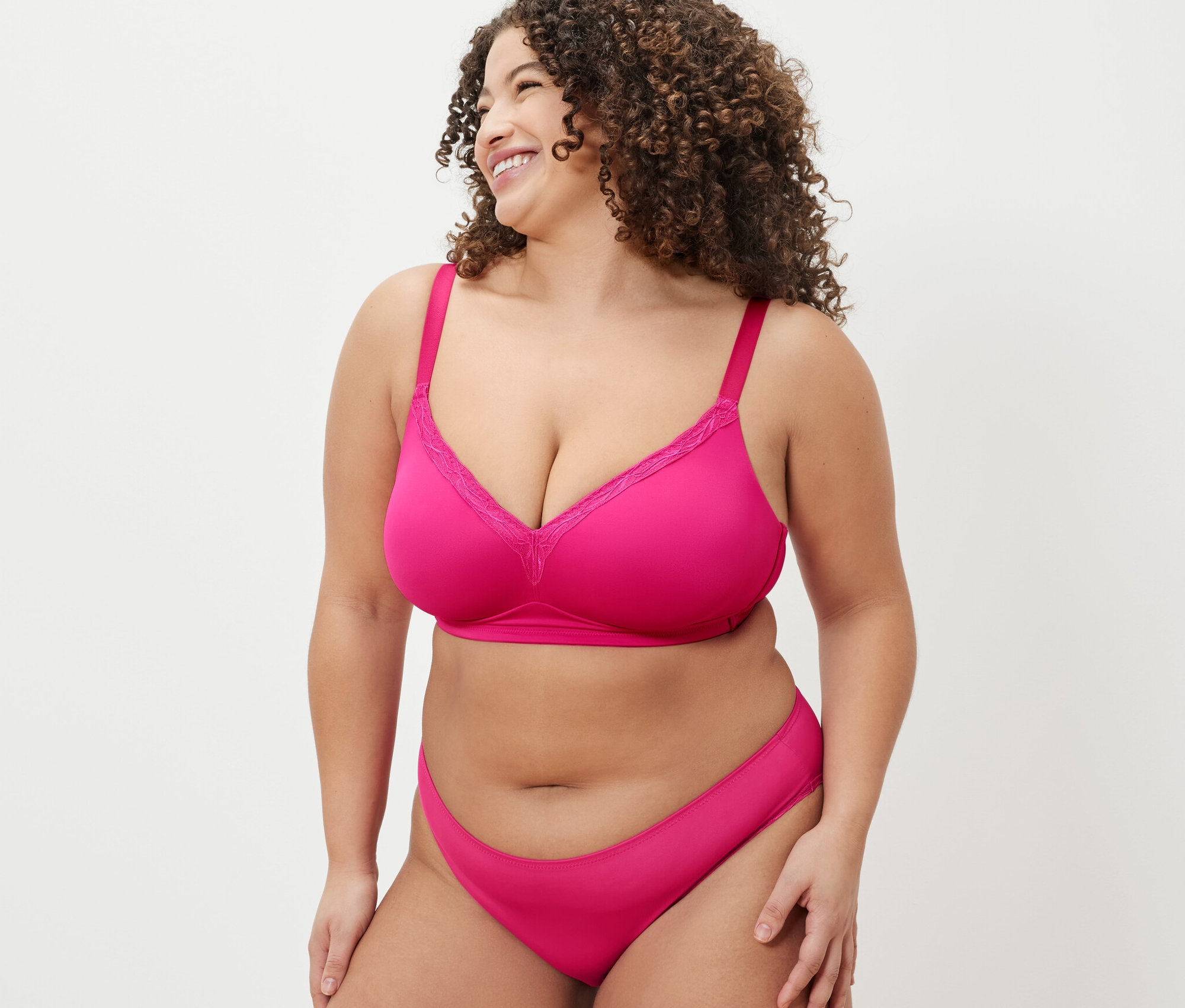 Kobieta z kręconymi włosami ma na sobie różowy biustonosz i majtki. Nosi miękki biustonosz i uśmiecha się.