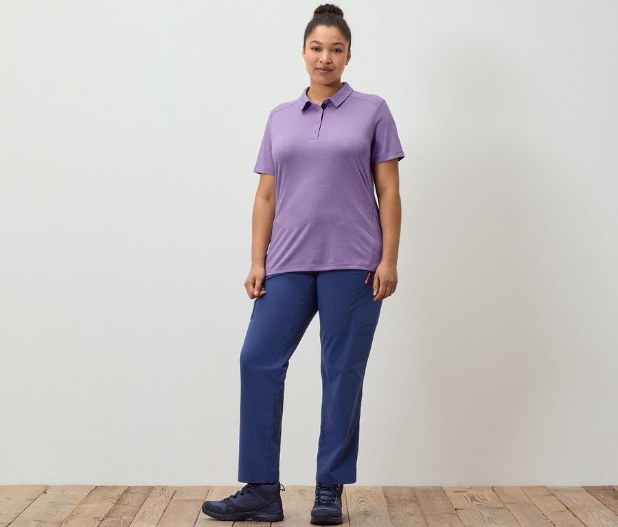 Kobieta pozuje na drewnianej podłodze w granatowych spodniach outdoorowych i fioletowej funkcjonalnej koszulce polo.