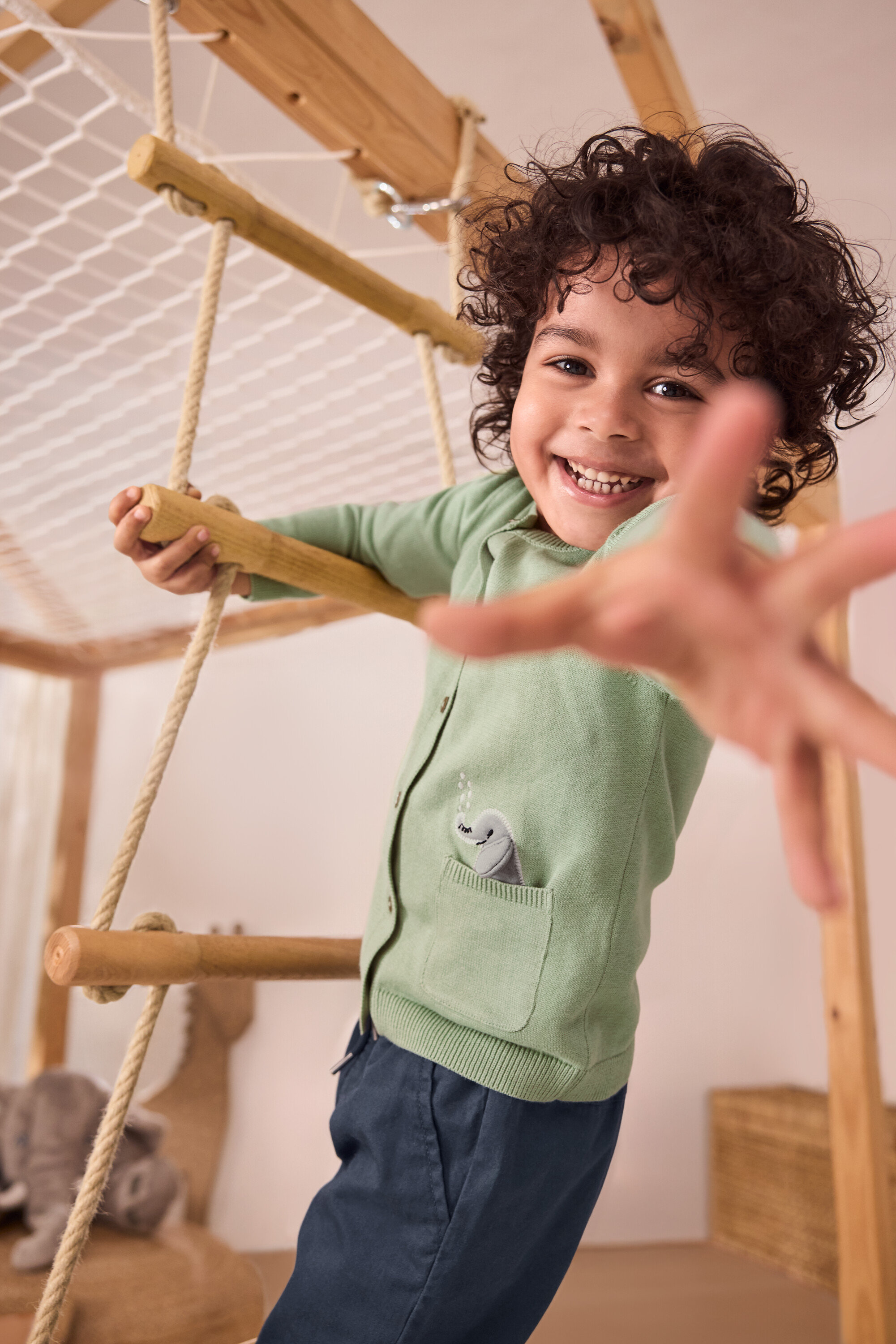 Uśmiechnięty chłopiec wspina się po drabince sznurkowej. Ma na sobie zielony sweter dziecięcy z aplikacją i niebieskie spodnie na gumce – fason „Anton”.